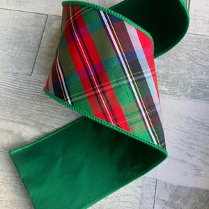 May include: A wide wired ribbon featuring a classic red, green, and white plaid pattern on one side and a solid deep green fabric on the reverse. The edges are finished with a matching green stitched border, allowing the ribbon to hold its shape.