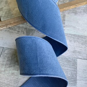 May include: A wide, dusty blue ribbon featuring a soft, velvet-like texture on the front side and a smooth, slightly shimmering finish on the reverse. The edges of the ribbon are finished with a matching stitched border for a clean, structured look.