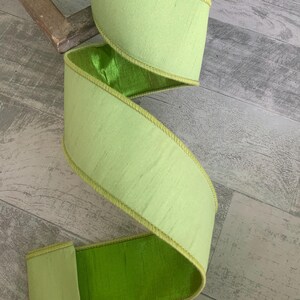 May include: A wide, decorative ribbon in a vibrant lime green color. The ribbon has a textured, silk-like appearance with a contrasting bright green interior. The edges are trimmed with a thin, yellow-gold stitching. The ribbon is likely used for crafting or decorating.