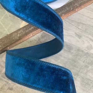 May include: A roll of teal velvet ribbon. The ribbon is thick and plush, with a soft, velvety texture. The edges are neatly stitched. The ribbon is partially unrolled, revealing the spool. The background is a light wood surface.