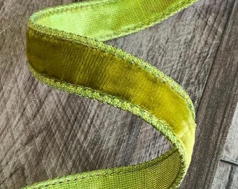 Puede incluir: Un trozo de cinta de terciopelo verde brillante con un centro texturizado y un borde cosido. La cinta está rizada y descansa sobre una superficie de madera. El color de la cinta es un verde amarillento vibrante.