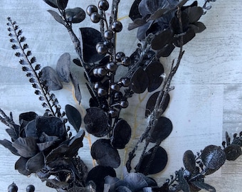 May include: A close-up of a black floral arrangement featuring various textures and shapes. The arrangement includes dark leaves, berries, and flower-like blooms, all in a deep black hue. Some elements have a glittery finish.