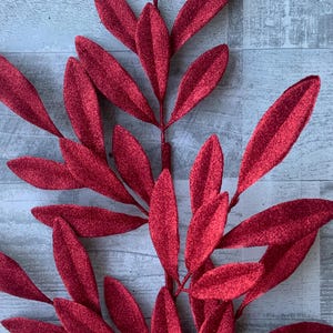 May include: A close-up of a decorative red glitter leaf stem. The leaves are elongated and have a textured, sparkling surface. The stem is a deep red color, and the leaves are arranged in a symmetrical pattern.