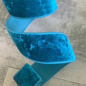 May include: A roll of teal velvet ribbon with a contrasting light blue satin backing. The ribbon is folded and curved, showcasing its plush texture and color variations. The ribbon is approximately 2 inches wide.