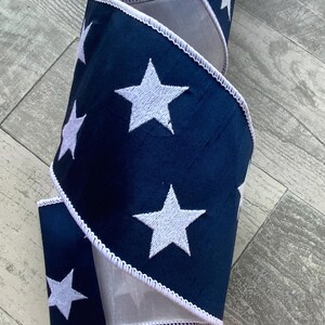 May include: Navy blue ribbon with white embroidered stars. The ribbon has a white scalloped edge and a gray backing. This decorative ribbon is suitable for crafts, gift wrapping, and holiday decorations.