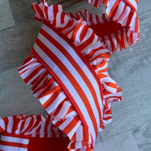 May include: A decorative ribbon featuring a vibrant orange and white striped pattern. The ribbon is designed with ruffled edges on both sides, creating a textured, layered appearance. The fabric displays a consistent vertical stripe motif throughout.