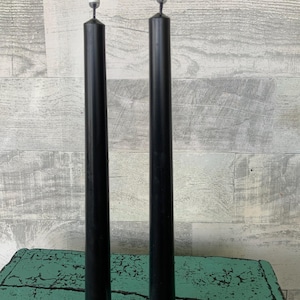 May include: Two black taper candles with white tips and bases stand upright. The candles are displayed against a weathered, light-colored wooden background. The candles are a classic, elegant design.