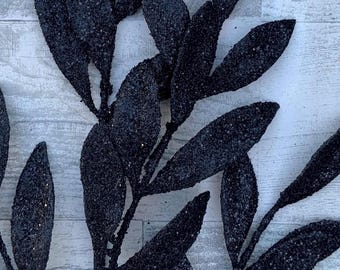 May include: Close-up of a decorative black leaf stem. The leaves are elongated and covered in glitter, creating a sparkling effect. The stem is dark and the background is a light, textured surface.