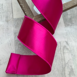 May include: A roll of vibrant fuchsia satin ribbon. The ribbon is thick and has a glossy sheen, with a visible stitched edge. The ribbon is curled and appears to be unwinding, showcasing its texture and color. Ideal for crafting and gift wrapping.