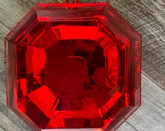 Könnte beinhalten: Ein roter, achteckiger, plastischer Weihnachtsbaumschmuck in Form eines großen, facettierten Edelsteins. Der Schmuck hat eine goldene Schnur, die oben befestigt ist.