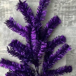 May include: A vibrant purple artificial Christmas tree branch. The branch features multiple offshoots with needle-like tinsel, creating a shimmering effect. The branch is a deep, rich purple color, perfect for holiday decorations.