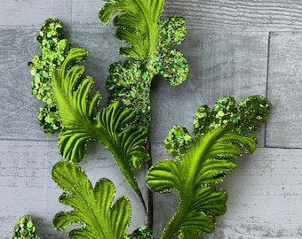 Puede incluir: Una rama de helecho artificial decorativa con hojas de color verde lima vibrante y una superficie texturizada y acanalada. Los bordes de las hojas están densamente adornados con lentejuelas verde brillante y purpurina fina, creando un acabado brillante.