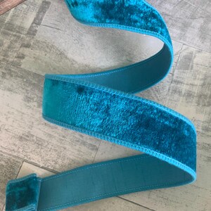 May include: A length of teal velvet ribbon with a contrasting teal satin backing. The ribbon is folded and curved, showing the plush texture of the velvet. The edges are neatly stitched with a lighter teal thread.