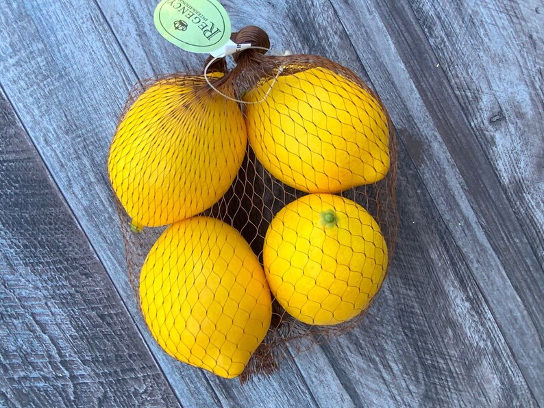 Puede incluir: Cuatro limones amarillos en una bolsa de malla marr&oacute;n. Los limones est&aacute;n dispuestos en c&iacute;rculo con la etiqueta de la bolsa visible. La etiqueta es verde y blanca con la palabra "Regent" impresa en ella.