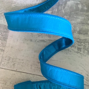 May include: A vibrant turquoise ribbon, likely satin, is displayed in a wavy pattern. The ribbon has a smooth, reflective surface and is edged with a contrasting white stitching. The ribbon's bright color and texture suggest it could be used for gift wrapping or crafting.
