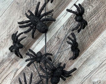 May include: A black Halloween decoration featuring multiple glittery spiders attached to a dark stem. The spiders have multiple legs and a fuzzy texture. The decoration is set against a wooden background.