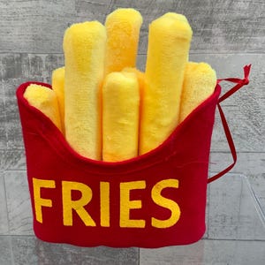 May include: A red plush toy shaped like a french fry container with yellow plush french fries inside. The container has the word "FRIES" printed on the front in yellow.