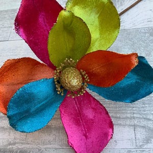 May include: A multi-colored velvet flower with a gold glitter center. The flower has petals in shades of pink, orange, green, and blue.