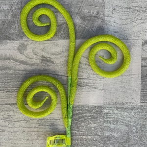 May include: A bright green decorative floral stem with three spiraling accents. The stem is wrapped in green fabric, and the spirals have a textured, glittery appearance. A yellow tag with a barcode is attached.