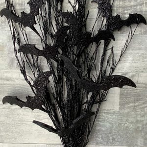 May include: Black glittery bat decorations attached to black branches. The bats are in various sizes and positions, creating a spooky Halloween display. The branches are arranged in a bundle, perfect for seasonal decor.