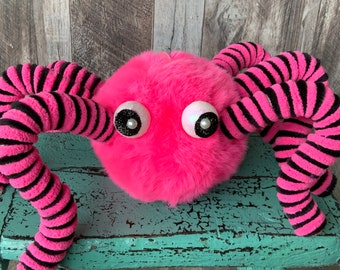 May include: A plush, pink creature with large, glittery eyes and black and pink striped tentacles. The toy has a fuzzy texture and a whimsical design, perfect for children's play or as a decorative item.