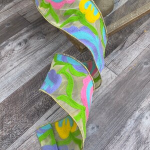 May include: A beige ribbon with embroidered flowers in shades of pink, yellow, blue, and green. The ribbon is curled and has a yellow border.