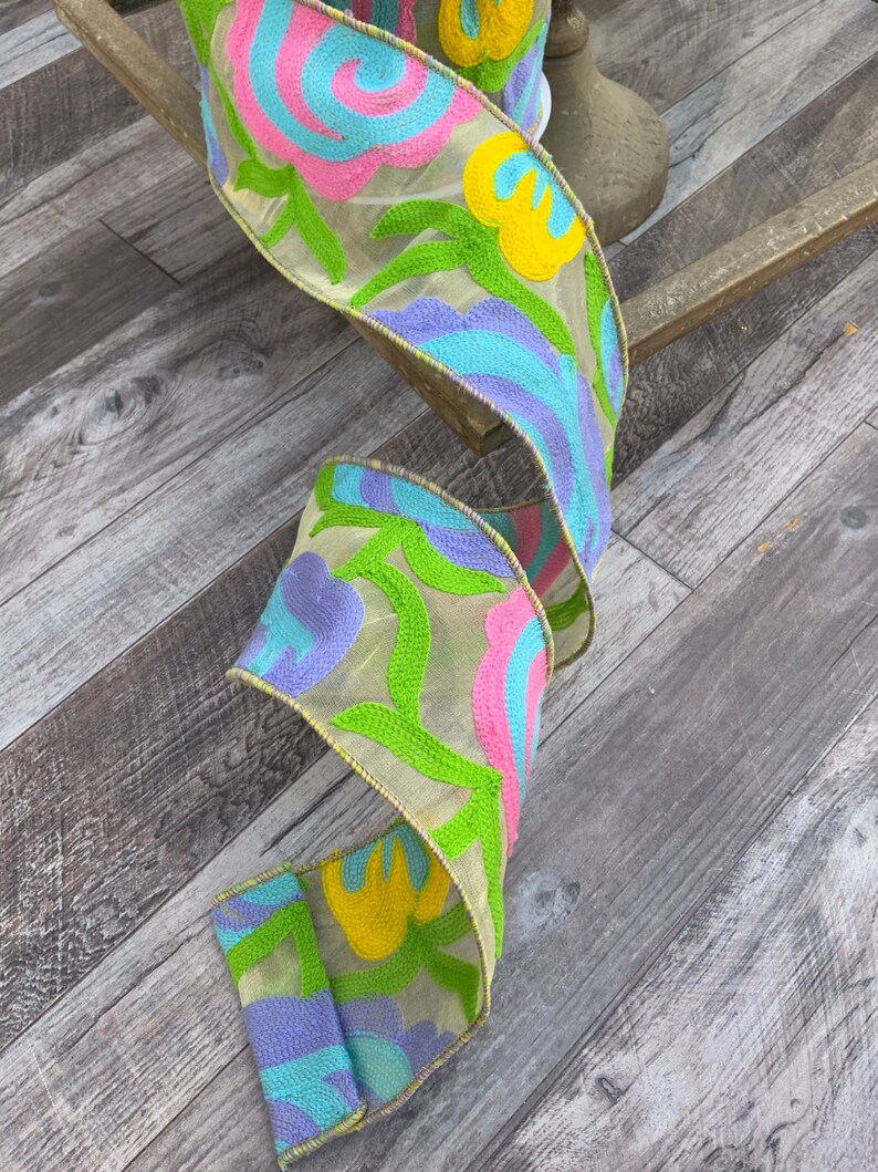 May include: A beige ribbon with embroidered flowers in shades of pink, yellow, blue, and green. The ribbon is curled and has a yellow border.