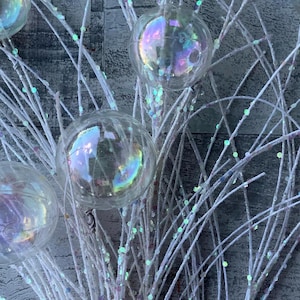 May include: Close-up of a decorative arrangement featuring white branches adorned with iridescent glass ball ornaments. The branches are speckled with iridescent glitter, creating a shimmering effect. The background is a textured gray surface.