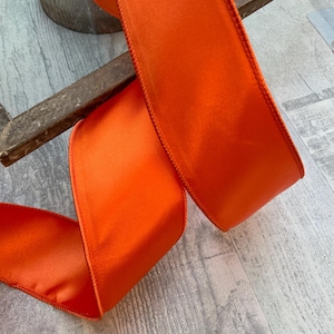 May include: Bright orange satin ribbon, with a stitched edge, is draped across a rustic wooden surface. The ribbon is folded and curved, showcasing its smooth texture and vibrant color. The ribbon is likely used for crafting or decorating.