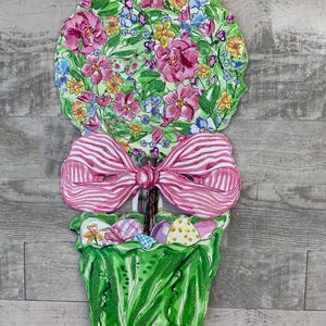 May include: A decorative Easter garden stake featuring a floral topiary design. The top is a circle of pink, blue, and yellow flowers on a green background. A pink and white striped bow sits above a green pot filled with Easter eggs.