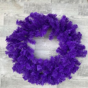 May include: A vibrant purple wreath with a circular shape. The wreath is made of artificial evergreen branches, creating a full and textured appearance. The bright purple color adds a bold and festive touch, suitable for seasonal or decorative use.