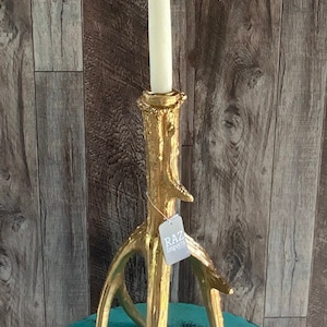May include: A gold-colored antler-shaped candle holder with a white candle. The candle holder has three legs and a textured surface. The tag reads "RAZ Imports".