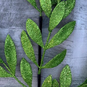 May include: A close-up of a decorative stem with glittery green leaves. The leaves are elongated and have a textured, sparkling surface. The stem is dark brown and appears to be wrapped. The background is a light gray, textured surface.