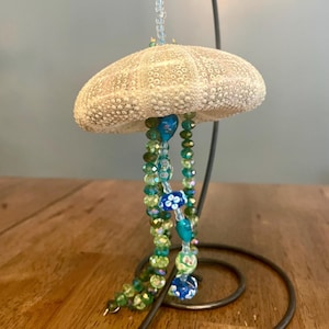 May include: A sea urchin ornament with green and blue beaded strands hanging from it. The ornament is hanging from a black metal stand.