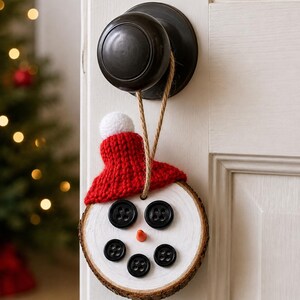 May include: A circular wood slice ornament painted white with a snowman face featuring five black buttons for eyes and a mouth, and a small orange cone nose. A red knitted hat with a white pom-pom sits on top, attached by a tan jute twine loop.