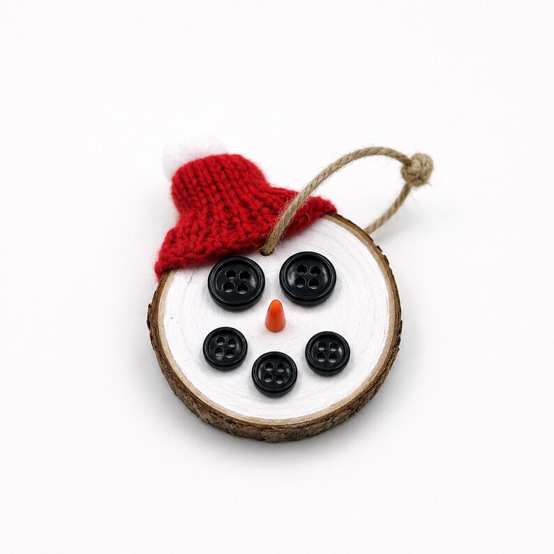 May include: A circular wood slice ornament painted white, featuring a face made of five black buttons and a small orange cone nose. A red knitted hat with a white pom-pom sits on top, and a jute twine loop is attached for hanging purposes.
