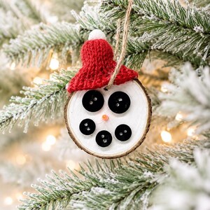 May include: A round wooden slice ornament painted white with a natural bark edge. It features five black buttons arranged to form eyes and a mouth, a small orange nose, and a red knitted hat with a white pom-pom on top, hanging by a twine loop.