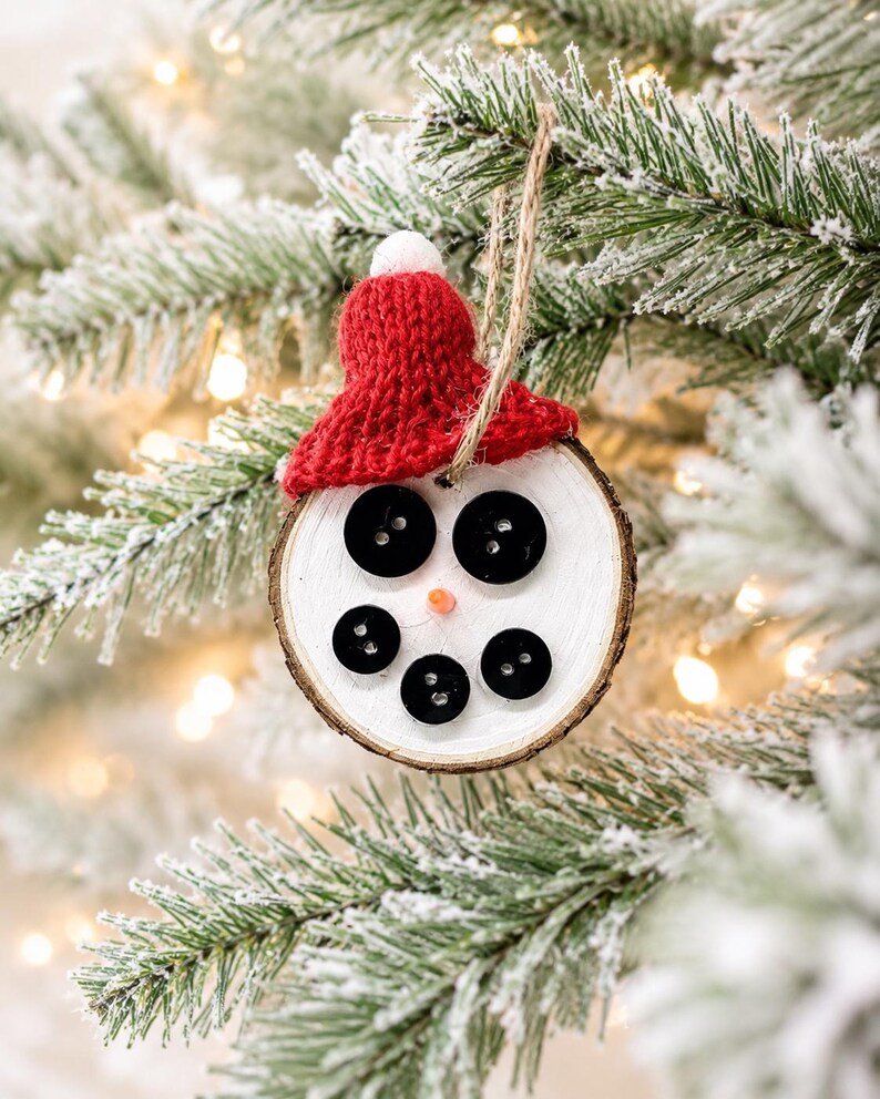 May include: A round wooden slice ornament painted white with a natural bark edge. It features five black buttons arranged to form eyes and a mouth, a small orange nose, and a red knitted hat with a white pom-pom on top, hanging by a twine loop.