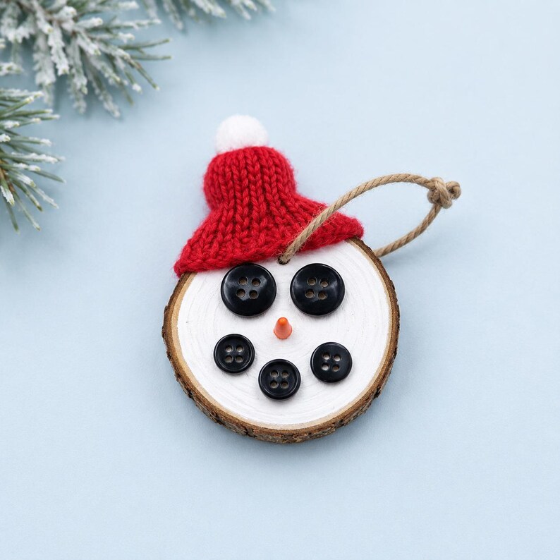 May include: A round wooden slice ornament painted white to resemble a snowman face. It features two black buttons for eyes, four black buttons for a mouth, and a small orange cone nose. A red knit hat with a white pom-pom sits on top with a jute string.