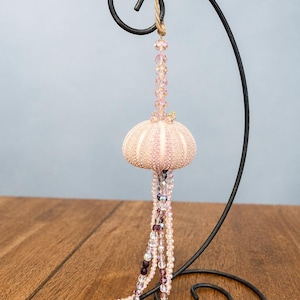 Op de afbeelding: Een hangend ornament met een lichtroze zee-egelschelp, bekroond met een verticale streng van gefacetteerde lichtroze kralen. Onder de schelp hangen meerdere strengen van doorzichtige, roze en donkerpaarse gefacetteerde kralen als tentakels.