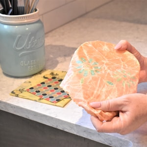 May include: A person is holding a peach-colored reusable food wrap with a green floral pattern. The wrap is made of beeswax, jojoba oil, and tree resin. It is a sustainable alternative to plastic wrap.