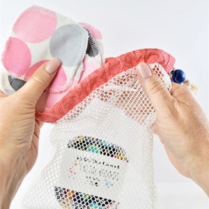 May include: A white mesh laundry bag with a pink and red drawstring top. The bag contains several round, pink and gray reusable makeup remover pads. The bag has a label that reads "Kiss Your Cotton" and "Facial Rounds".