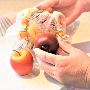 May include: A white mesh produce bag with a colorful floral pattern and drawstring closure. Two apples, one red and one yellow, are inside the bag.