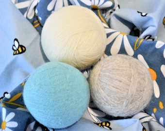 May include: Three wool dryer balls in different colors: beige, light blue, and gray. The balls are on a blue and white fabric with a daisy pattern.
