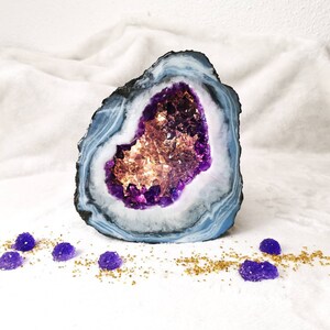 May include: A large amethyst geode with a purple and white interior. The geode is sitting on a white surface with gold glitter and purple candies.