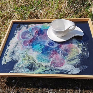 Puede incluir: Una bandeja de servir de madera con una tapa de resina negra con remolinos coloridos abstractos y una taza y un platillo blancos.