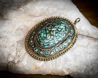 May include: Oval pendant with a mosaic of turquoise stones set in a silver and gold-toned metal frame. The pendant has a loop for a chain and is displayed on a white, textured surface.