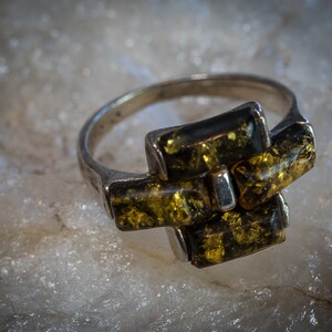 May include: A silver ring with a green and gold amber stone setting. The stone is cut into multiple rectangular shapes and is set in a geometric pattern.