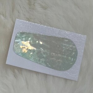 Può includere: Una carta bianca con un adesivo olografico con un design astratto blu chiaro, verde e argento.