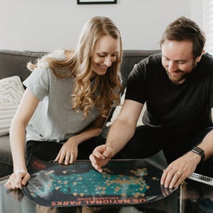 May include: A couple sitting on a sofa, looking at a map of the United States National Parks. The map is spread out on a glass coffee table. The map is labelled "US National Parks Explorer's Guide".